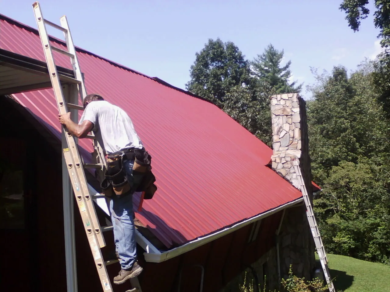 Roofer on a ladder beside a red metal roof and stone chimney — Soffit & Fascia Repair by Roofing Brothers in Earlville